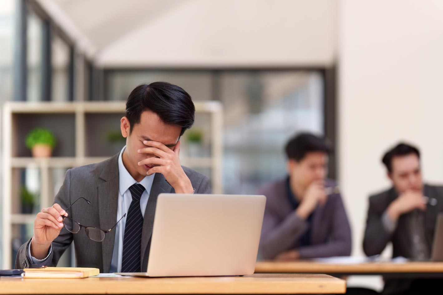 一位穿著正式西裝的男性在辦公室環境中顯得沮喪和疲憊，手扶著額頭，眼鏡拿在另一手上，看起來像是在長時間的工作後感到壓力，電腦筆記本放在桌子上，背景中有模糊的同事正在交談。