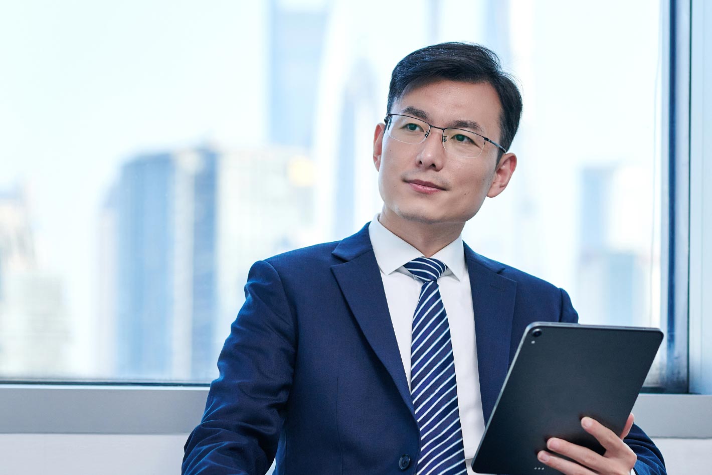 一位亞洲男士穿著正式的藍色西裝、條紋領帶，帶著眼鏡，專業而自信地在辦公室環境中看著攝像頭，手持平板電腦。